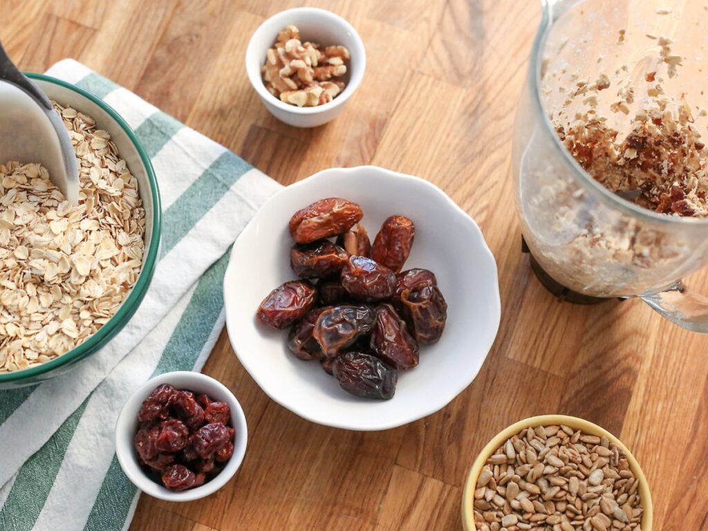 oatmeal dates seeds nuts in bowls with a measuring scoop and a pitcher on a wood table